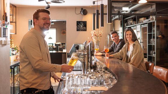 Ein freundlicher Barkeeper serviert ein Getränk an der Bar. Im Hintergrund sitzen zwei fröhliche Gäste und genießen ihre Zeit. - Bäckelar Wirt - Sölden
