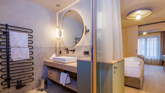 Ein modernes Badezimmer mit einem runden Spiegel und einer stilvollen Waschbeckenanlage. Im Hintergrund ist ein gemütliches Schlafzimmer mit Fenster zu sehen. - Bäckelar Wirt - Sölden