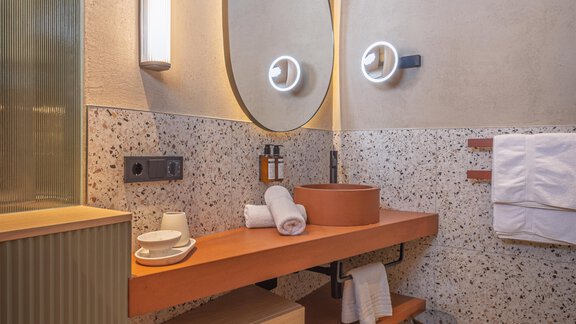 Ein modernes Badezimmer mit einer stilvollen Holzregal. An der Wand befinden sich ein runder Spiegel und eine dekorative Beleuchtung. - Bäckelar Wirt - Sölden
