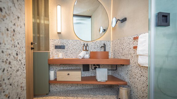 Ein modernes Badezimmer mit einem runden Spiegel und Holzregalen. Die Wände sind mit dekorativem Material gestaltet und es gibt frische Handtücher. - Bäckelar Wirt - Sölden