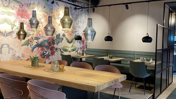 Ein modernes Restaurant mit einer großen Holztisch und stilvollen Stühlen. Die Wände sind mit bunten Blumenmotiven dekoriert, und es hängen elegante Lampen von der Decke. - Bäckelar Wirt - Sölden