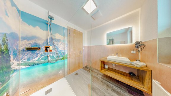 A modern bathroom with a clear shower cubicle and a wooden vanity. In the background, a beautiful landscape featuring mountains and water is depicted. - Belmonte Sölden - Sölden
