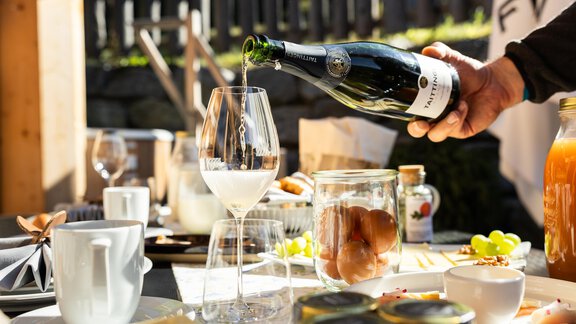 A set table with various foods and drinks. A person is pouring sparkling wine into a wine glass. - Chalet Resort Sölden - Sölden
