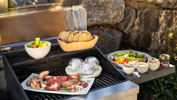 A grill with various types of meat, potatoes, and a salad bowl. Next to it are fresh vegetables and bread. - Chalet Resort Sölden - Sölden