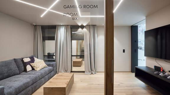 A modern gaming room with a cozy sofa and a wooden table. The walls are painted in neutral colors, and there is a large TV setup. - Chalet White Pearl by MYALPS - Sölden