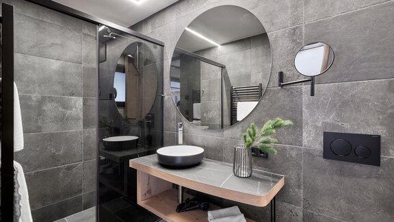 A modern bathroom with gray tiles and elegant fixtures. There is a shower, a round mirror, and a stylish vanity. - Chalet White Pearl by MYALPS - Sölden