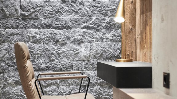 A modern workspace with a stylish chair and a simple desk. The wall is made of gray stone and complements the wooden paneling well. - Chalet White Pearl by MYALPS - Sölden