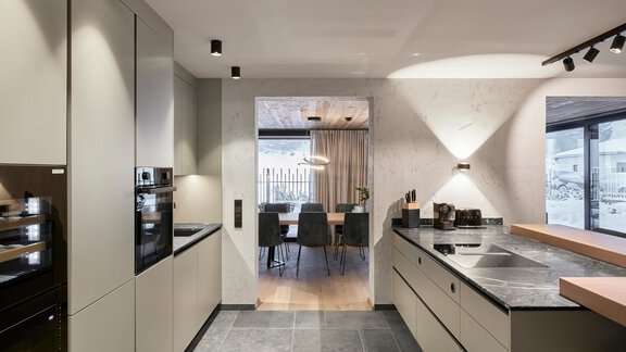 A modern kitchen with elegant gray cabinets and high-quality countertops. In the background, a dining area with a large table and chairs is visible. - Chalet White Pearl by MYALPS - Sölden