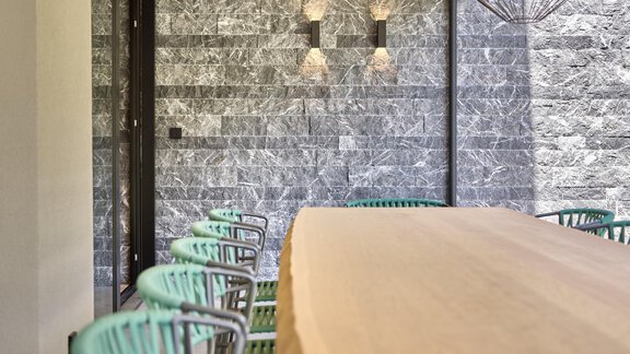 A modern dining area with a long wooden table and green chairs. The walls are made of gray stone and the lighting is stylish. - Chalet White Pearl by MYALPS - Sölden