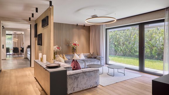 A modern furnished living room with a comfortable sofa and large windows. The room design is bright and inviting, with a view of a garden. - Chalet White Pearl by MYALPS - Sölden