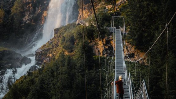 A suspension bridge leads over a gorge with a waterfall in the background. Green trees surround the scene, giving it a natural beauty. - Chalet White Pearl by MYALPS - Sölden