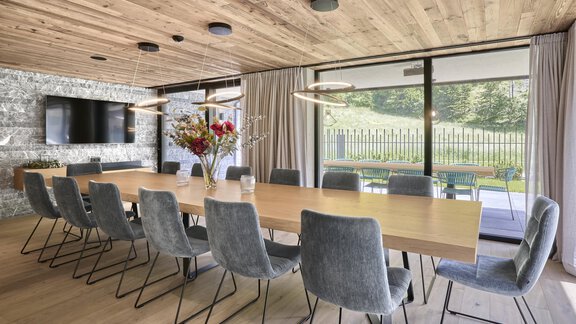 A modern conference room with a large wooden table and gray chairs. Large windows provide a view of the green surroundings and lots of natural light. - Chalet White Pearl by MYALPS - Sölden