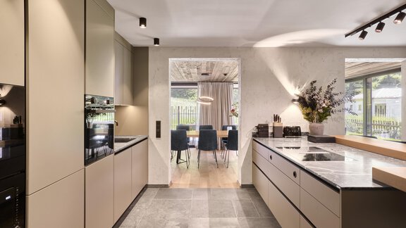 A modern kitchen with elegant beige cabinets and a large, bright window. In the background, there is a dining area with stylish chairs and a table. - Chalet White Pearl by MYALPS - Sölden