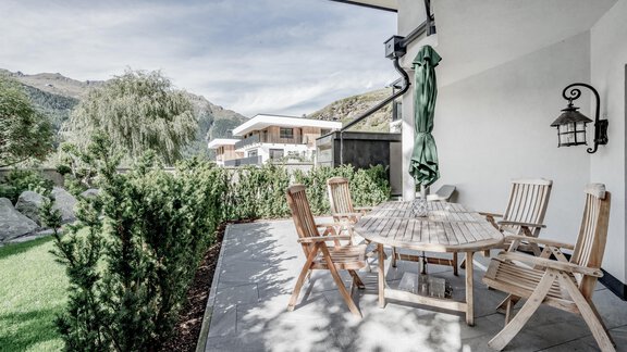 A cozy outdoor area with a round table and wooden chairs. The surroundings are green with plants and a view of the mountains. - CORSO living Aparthotel - Sölden