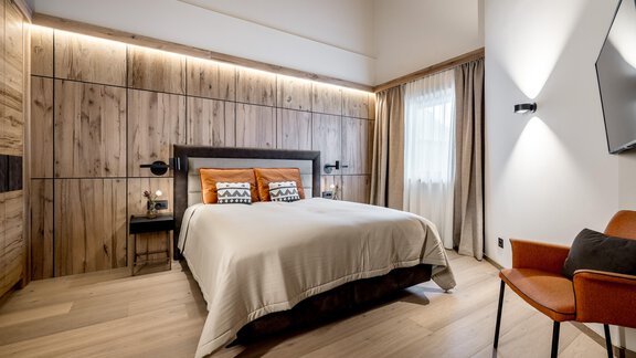 Ein modernes Schlafzimmer mit Holzverkleidungen und sanfter Beleuchtung. Ein Bett mit orangefarbenen Kissen und ein Stuhl ergänzen die elegante Einrichtung. - Das Central - Alpine . Luxury . Life - Sölden