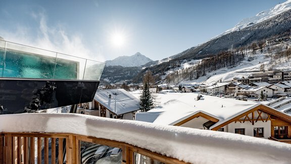 Eine verschneite Landschaft mit Bergen im Hintergrund und einem klaren blauen Himmel. Im Vordergrund befindet sich ein Balkon mit Holzgeländer. - Das Central - Alpine . Luxury . Life - Sölden