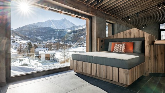 Ein modernes Schlafzimmer mit einem großen Fenster und Blick auf schneebedeckte Berge. Die Sonne scheint und es gibt eine gemütliche Liege mit bunten Kissen. - Das Central - Alpine . Luxury . Life - Sölden