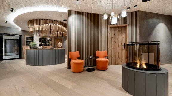 Ein modernes Empfangsbereich mit warmen Holztönen und stilvollen Möbeln. Zwei orangefarbene Stühle stehen neben einem dekorativen Kamin. - Das Central - Alpine . Luxury . Life - Sölden
