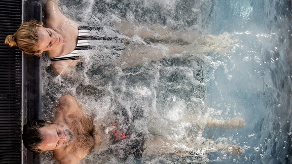 Zwei Personen entspannen im Wasser eines Whirlpools. Spritzendes Wasser umgibt sie, während sie die Zeit genießen. - Das Central - Alpine . Luxury . Life - Sölden