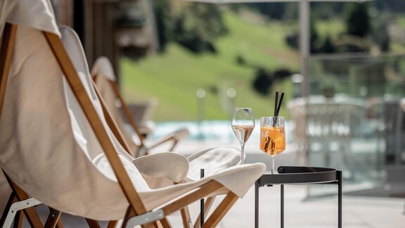 Ein entspannter Bereich mit Liegestühlen und zwei Gläsern Getränken auf einem Tisch. Im Hintergrund ist eine grüne Landschaft und ein Swimmingpool sichtbar. - Das Central - Alpine . Luxury . Life - Sölden