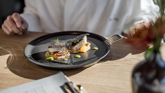 Ein kunstvoll angerichteter Teller mit Fisch und bunten Saucen. Im Hintergrund ist eine gemütliche Tischatmosphäre zu erkennen. - Das Central - Alpine . Luxury . Life - Sölden