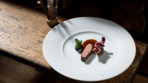 Ein kunstvoll angerichteter Teller mit zartem Fleisch, buntem Gemüse und einer Sauce. Der Tisch ist aus Holz und hat eine rustikale Ausstrahlung. - Das Central - Alpine . Luxury . Life - Sölden