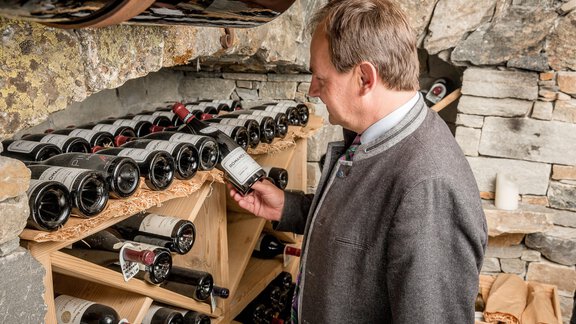Ein Mann schaut sich eine Flasche Wein in einem Weinkeller an. Im Hintergrund sind viele Weinflaschen auf Regalen angeordnet. - Das Central - Alpine . Luxury . Life - Sölden