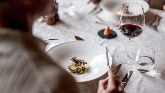 Ein elegantes Gericht auf einem Tisch, umgeben von Besteck und Gläsern. Im Hintergrund wird ein Glas Rotwein serviert. - Das Central - Alpine . Luxury . Life - Sölden