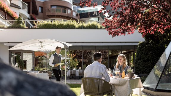 Ein schönes Restaurant im Freien mit einem Tisch für zwei Personen unter einem Sonnenschirm. Ein Kellner serviert Getränke in einer angenehmen Umgebung. - Das Central - Alpine . Luxury . Life - Sölden