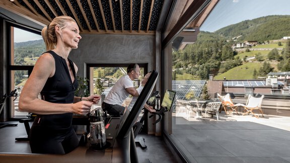 Eine Frau trainiert an einem Laufband in einem modernen Fitnessstudio mit großen Fenstern. Im Hintergrund sind grüne Hügel und eine Sonnenterrasse sichtbar. - Das Central - Alpine . Luxury . Life - Sölden