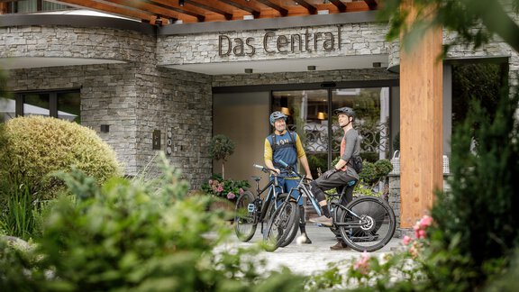 Zwei Radfahrer stehen vor dem Hotel "Das Central". Die Umgebung ist grün und einladend, mit einer modernen Architektur im Hintergrund. - Das Central - Alpine . Luxury . Life - Sölden