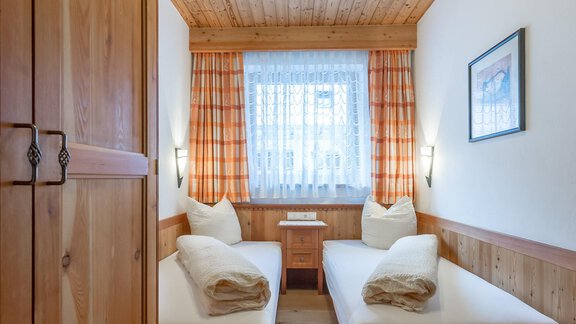 A cozy bedroom with two single beds and wooden paneling. The room has large windows with curtains and a warm, inviting atmosphere. - Ferienappartements Linserhof - Sölden