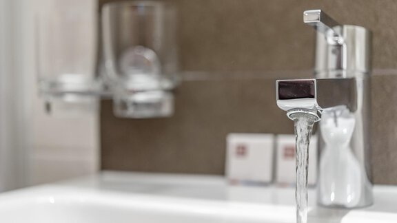 A modern faucet with flowing water in an elegant bathroom. In the background, a glass and toiletries can be seen. - Ferienhaus Apart Sonnzeit - Sölden