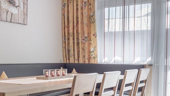 A modern dining area with a long wooden table and bright chairs. Curtains and a glass window create an inviting atmosphere. - Ferienhaus Apart Sonnzeit - Sölden