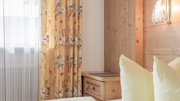 A cozy bedroom with bright, floral curtains and a wooden wall. On the bed are large, yellow cushions. - Ferienhaus Apart Sonnzeit - Sölden