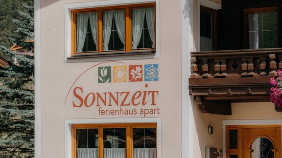 A holiday home named "Sonnzeit" is attached to the wall. The facade has large windows with white curtains and a rustic balcony cladding. - Ferienhaus Apart Sonnzeit - Sölden