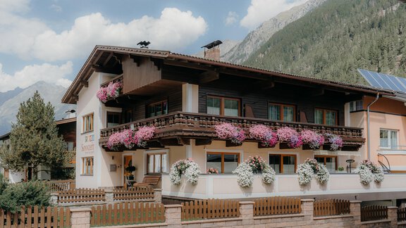 A charming house with balconies adorned with colorful flowers. In the background, mountains and a blue sky are visible. - Ferienhaus Apart Sonnzeit - Sölden
