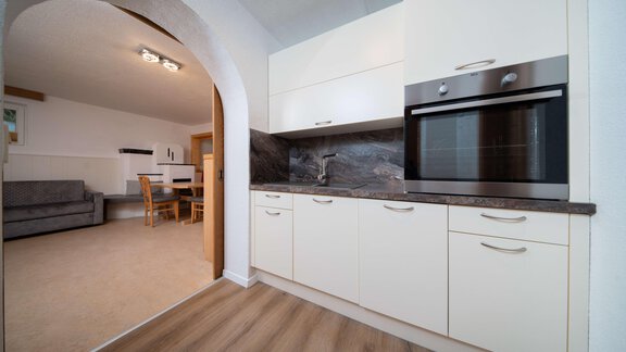 A modern kitchen with white cabinets and an oven. In the background, a cozy living area is visible. - Ferienhaus F2 - Sölden - Sölden