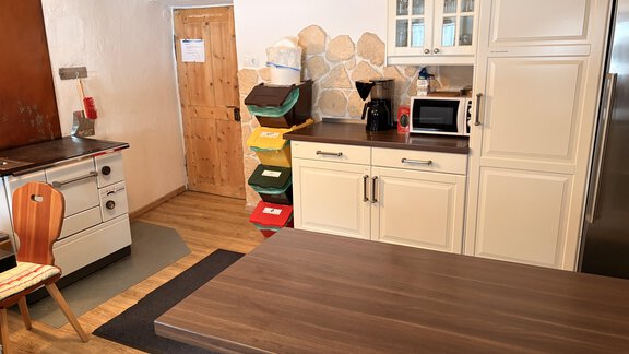 A modern kitchen with a wooden table, modern appliances, and an adjacent living area. The walls are light-colored, and there are various recycling bins. - Ferienhaus Kaspers - Sölden