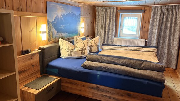 A cozy bedroom with a large bed and wooden-paneled walls. The decoration includes cushions with a antler pattern and a beautiful mountain picture on the wall. - Ferienhaus Kaspers - Sölden