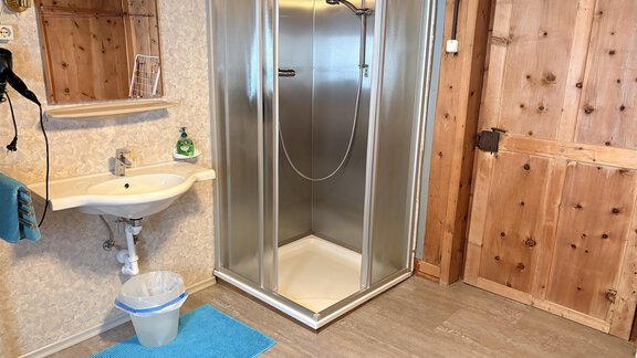 A simple bathroom with a shower cubicle and a sink. The walls are adorned with wood and wallpaper, and there is a blue bath mat on the floor. - Ferienhaus Kaspers - Sölden