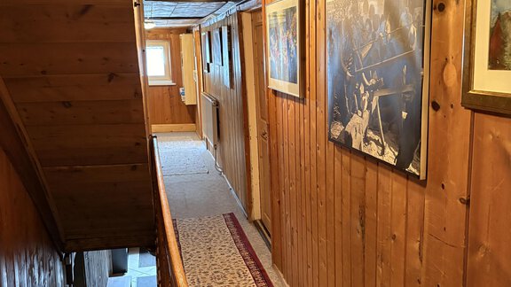 A long hallway with wood paneling and a patterned carpet. Various pictures hang on the walls, and a ceiling lamp provides light. - Ferienhaus Kaspers - Sölden