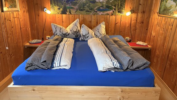 A cozy bedroom with a large bed and blue bed sheets. The walls are made of wood and there are pictures of mountains in the background. - Ferienhaus Kaspers - Sölden