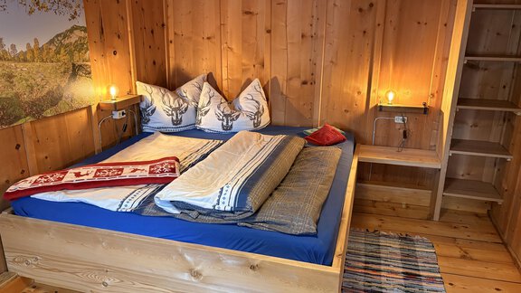 A cozy bedroom with a wooden double bed and colorful cushions. The walls are made of wood, and there is a corner with a small dresser. - Ferienhaus Kaspers - Sölden
