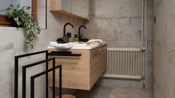 A modern bathroom with a wooden washbasin and black fittings. The walls are designed in a neutral concrete style, and there is a small plant decoration. - Ferienhaus Valsora - Sölden