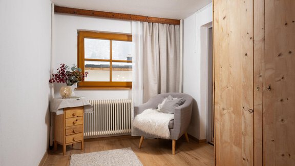 A cozy room with a gray armchair, a wooden table, and a large wardrobe. The window lets in a lot of light and the decoration is simple and modern. - Ferienhaus Valsora - Sölden