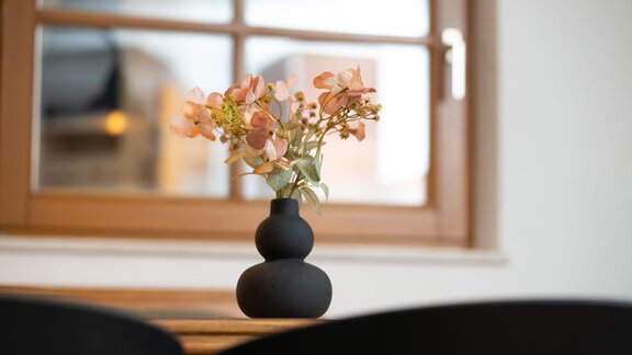 A black vase with artificial, bright flowers stands on a table. In the background, a window with brown frames is visible. - Ferienhaus Valsora - Sölden