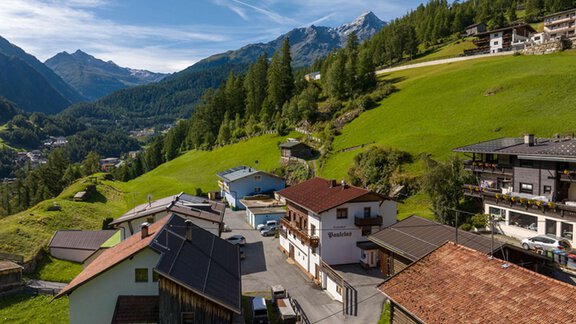 A picturesque mountain landscape with green meadows and snow-covered peaks. In the foreground, traditional houses and a quiet road can be seen. - Ferienhof Paulelas - Sölden