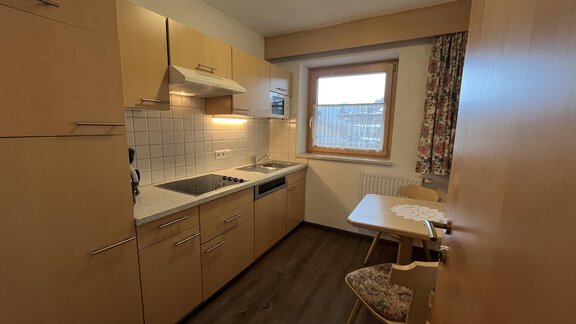 A cozy kitchen with wooden cabinets and modern appliances. A small table stands next to the window with a view outside. - Franzele Appartements & Zimmer - Sölden