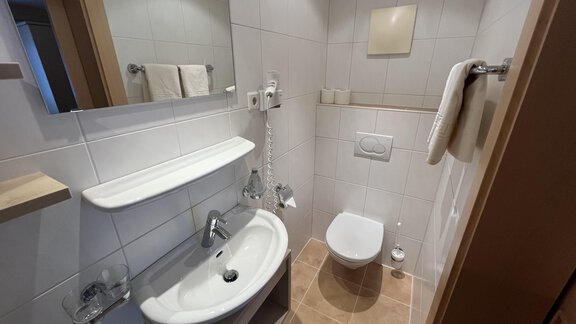 A modern bathroom with white tiles and a bright sink. It includes a toilet, towels, and a large mirror. - Franzele Appartements & Zimmer - Sölden
